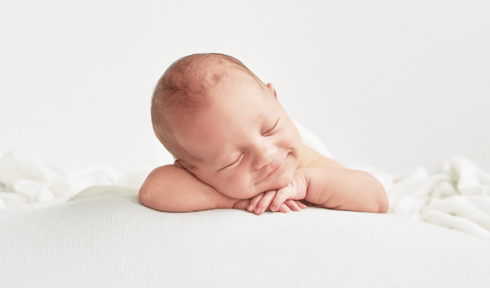 foto de uma bebê de poucos meses deitado de bruços, de olhos fechados e sorrindo